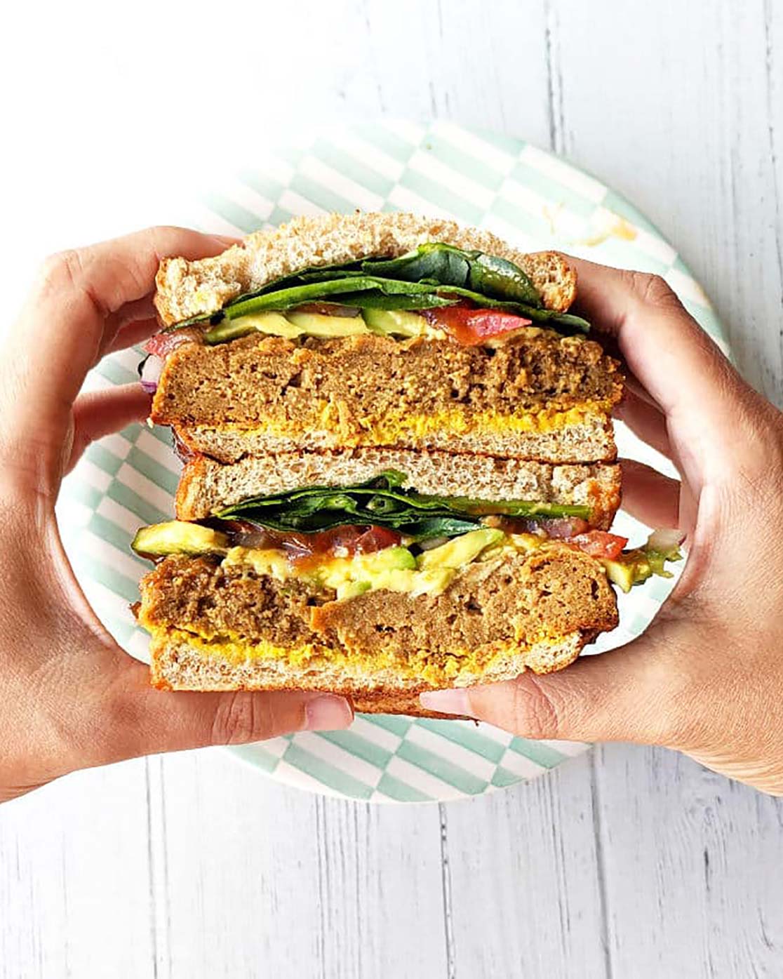 Hands holding a stacked vegan meat and salad sandwich.