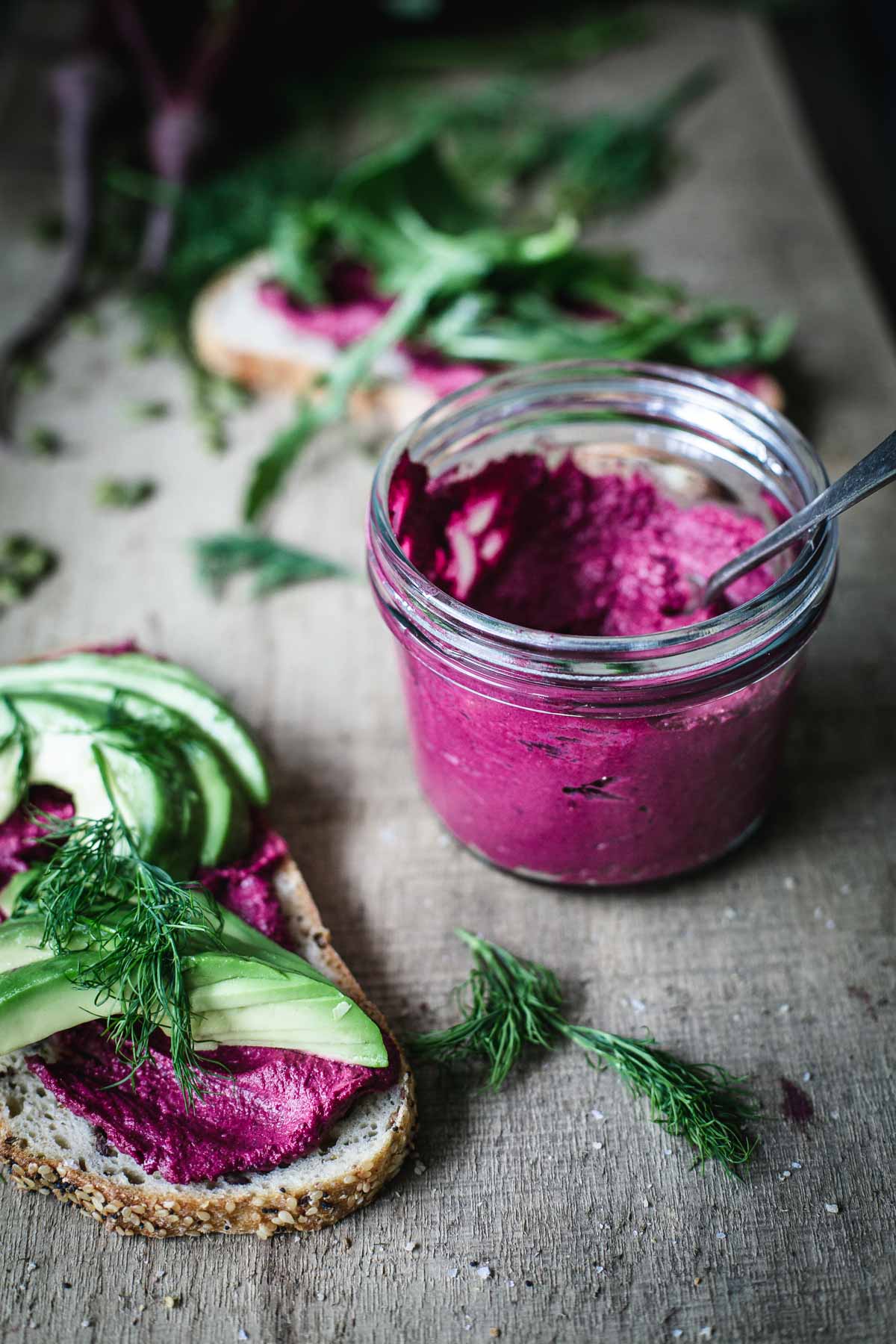 A jar of beetroot humus with bread nearby on a wooden board.