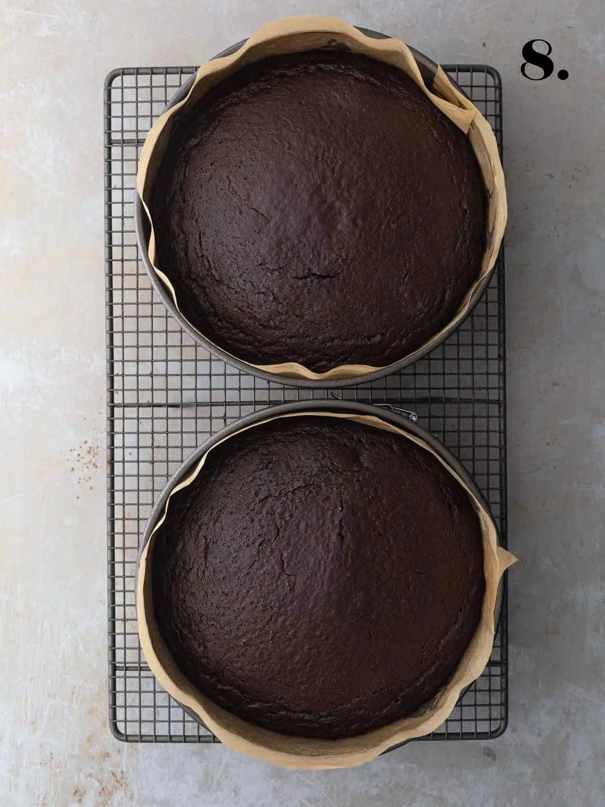 Two freshly baked round vegan chocolate cakes in their baking pans on a wire cooling rack.