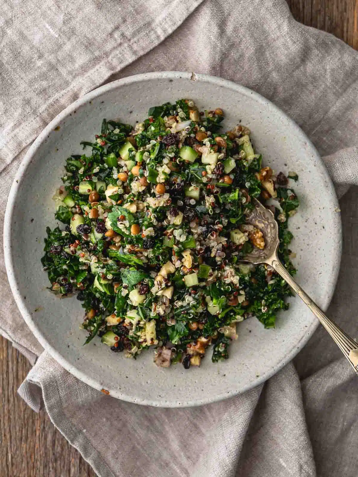A wooden table with a bowl of kale and quinoa salad with a spoon and linen napkin.